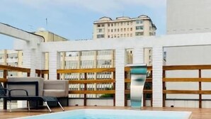 Una piscina con muelle de madera, sillones de descanso y un edificio con balcones al fondo.
