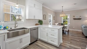 A modern kitchen with white cabinetry, a stainless steel sink, and a dishwasher. There is a dining area with a table and chairs, and a gray sofa.