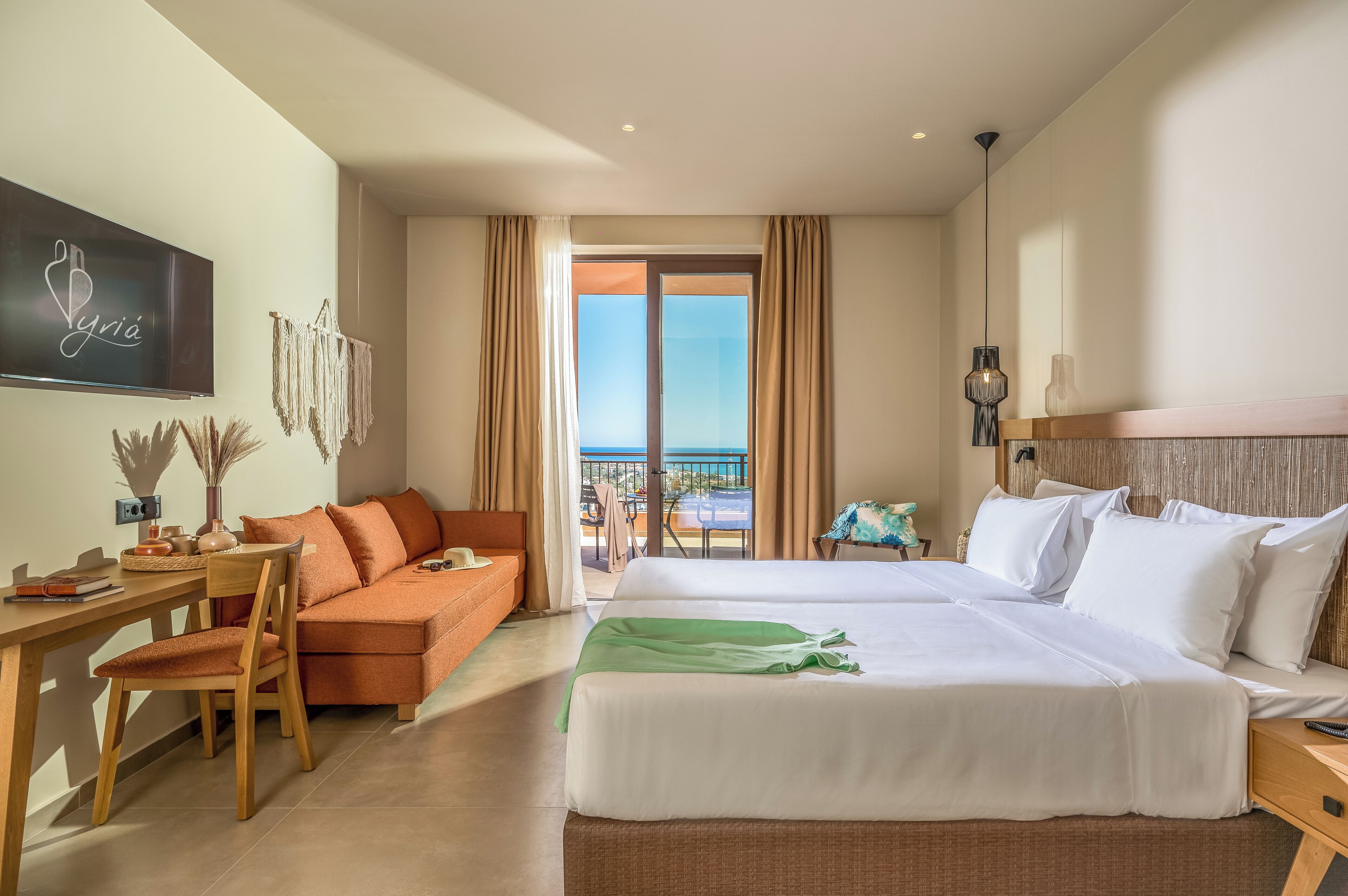 A hotel room with a large bed, a desk with a chair, a sofa, and a view of the sea through a glass door.