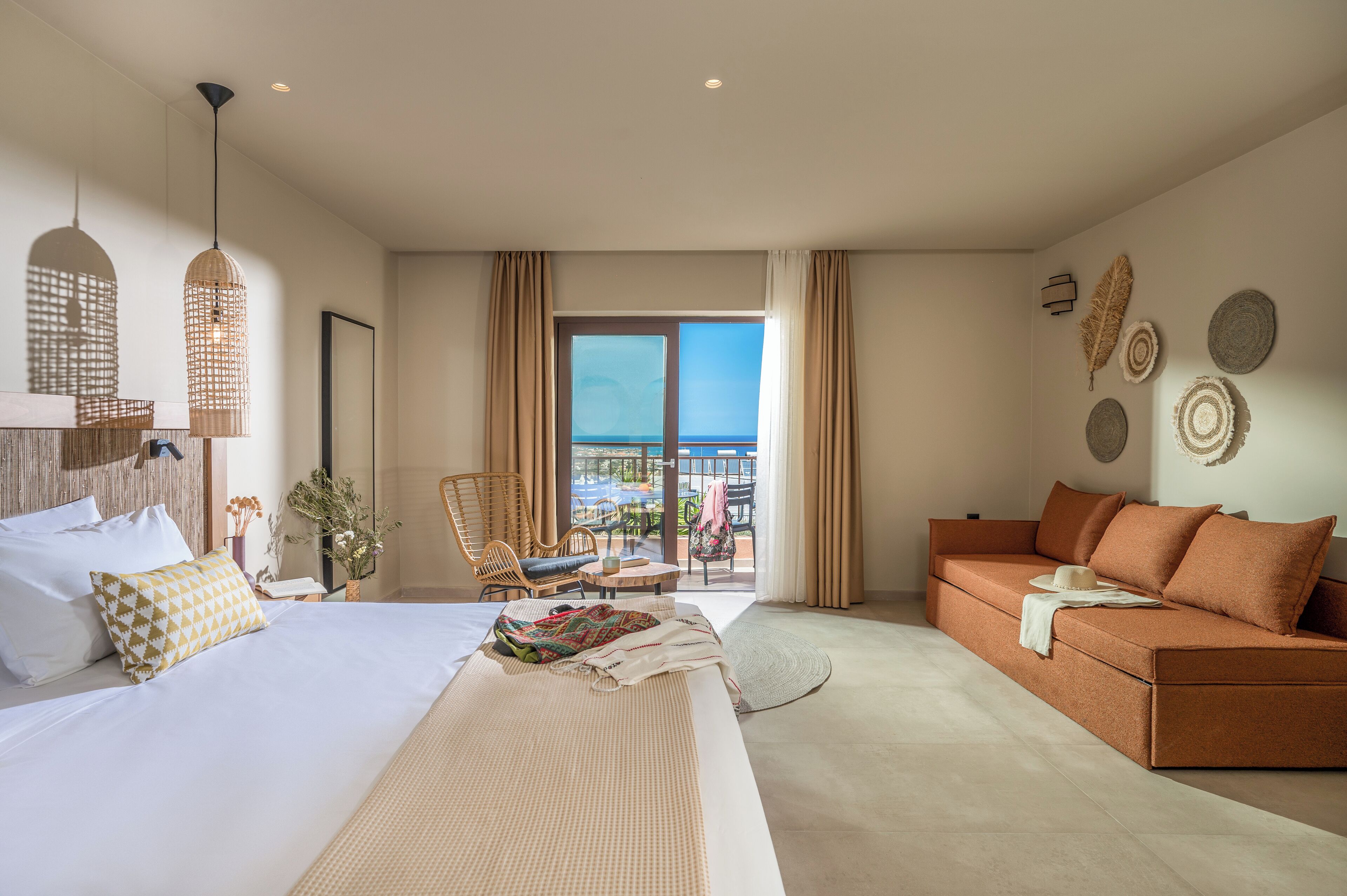 A hotel room with a bed, a sofa, a dining table, and a balcony with a view of the sea.