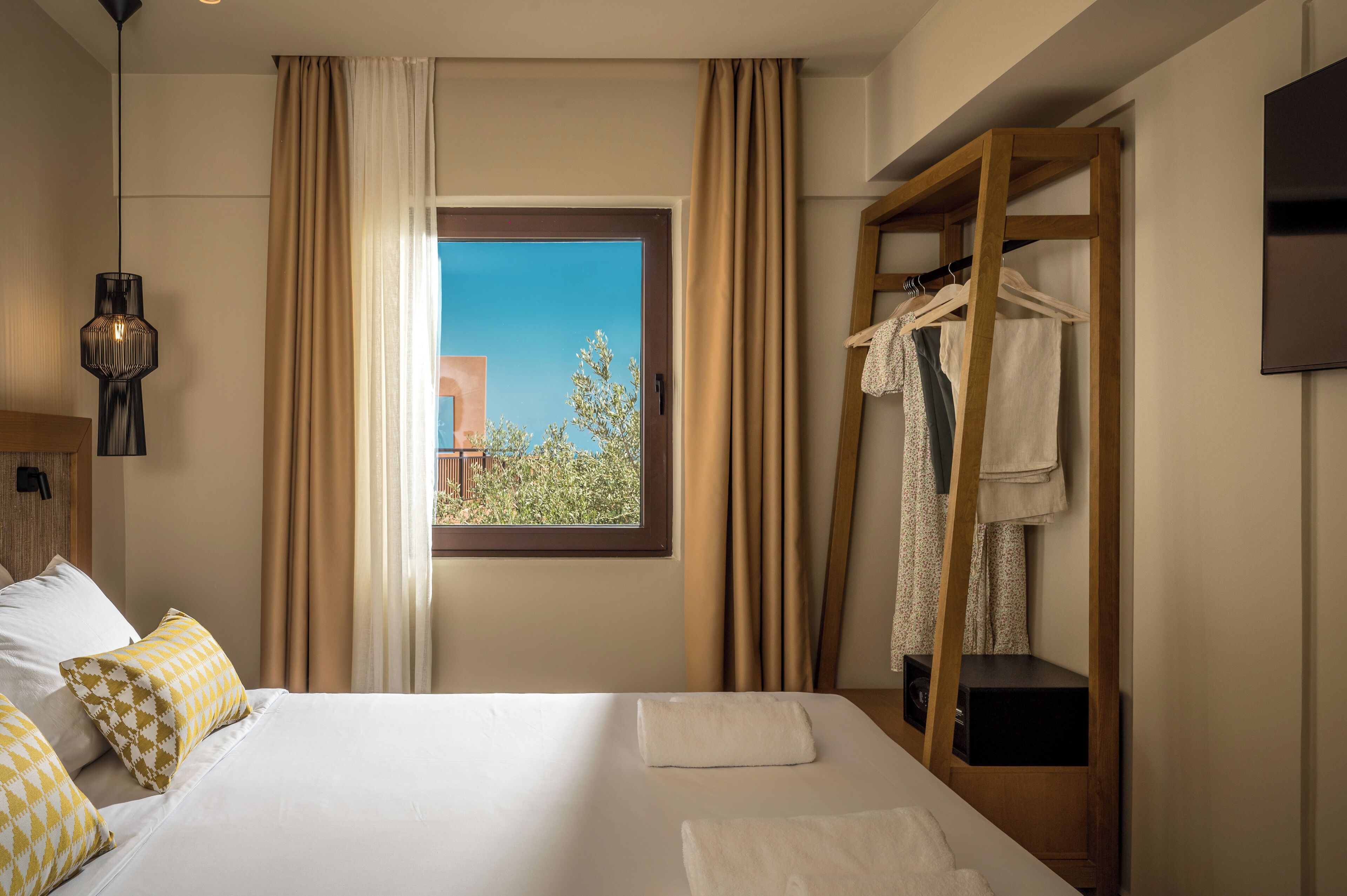 A hotel room with a bed, a window with curtains, a hanging clothes rack, and a pendant light.