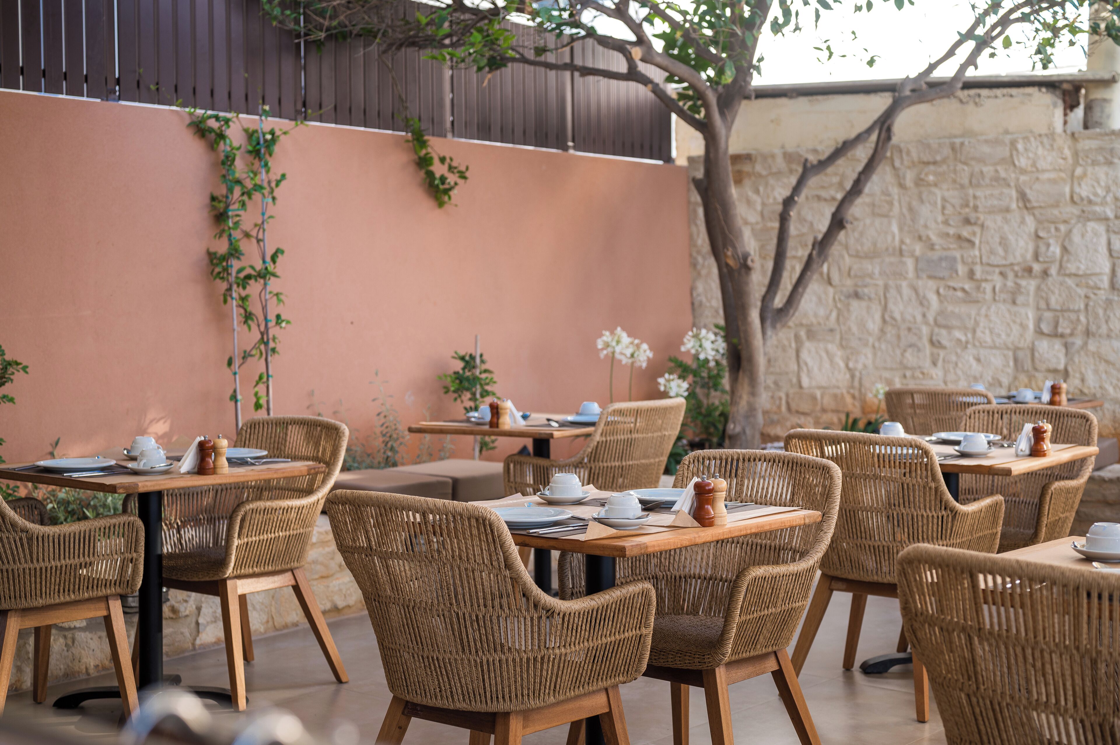 An outdoor dining area with wicker chairs and tables, set against a stone wall and a wooden fence.