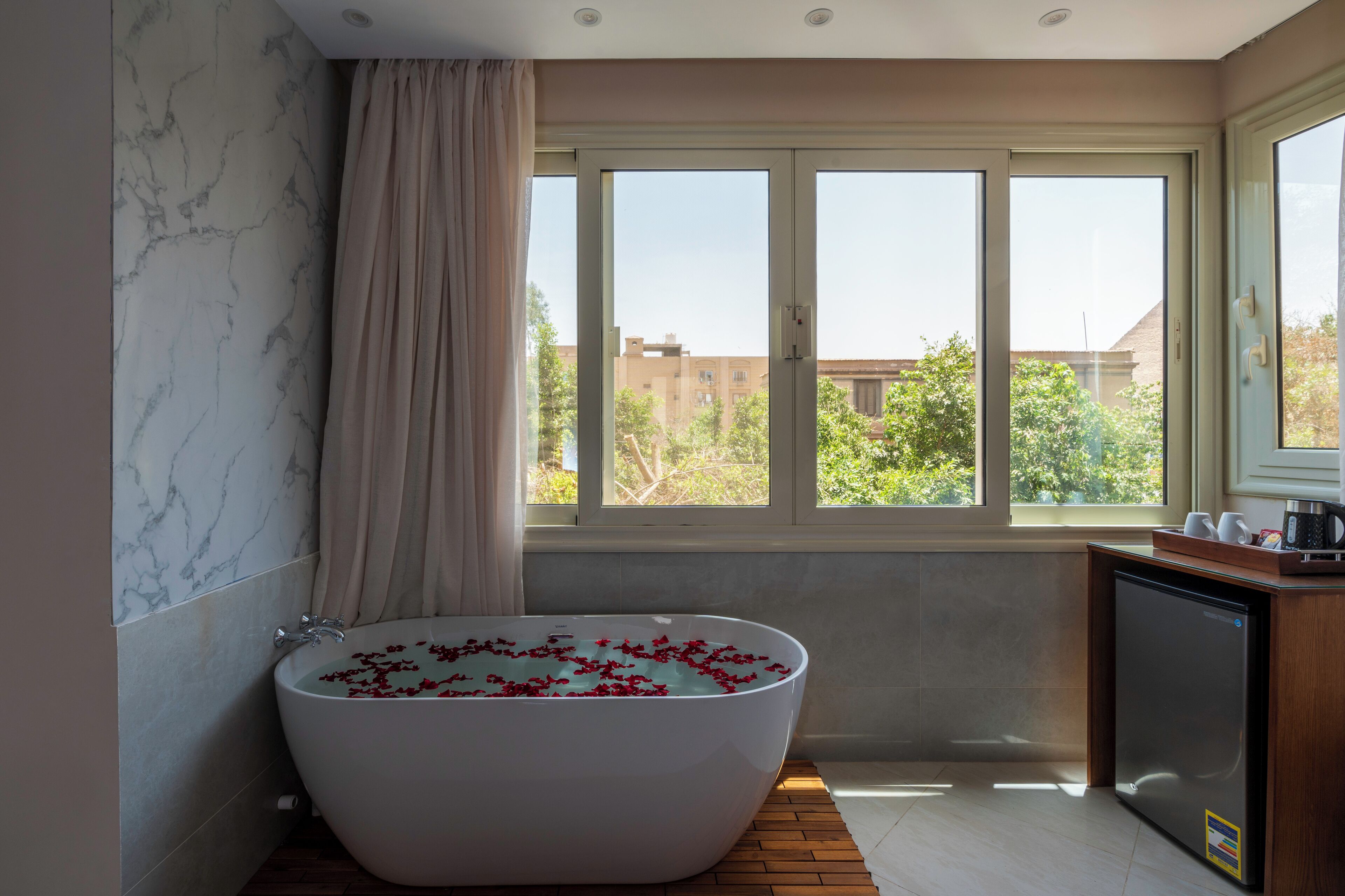 A modern bathroom with a freestanding bathtub filled with rose petals, a window with a view of buildings and trees, and a small refrigerator on the right.