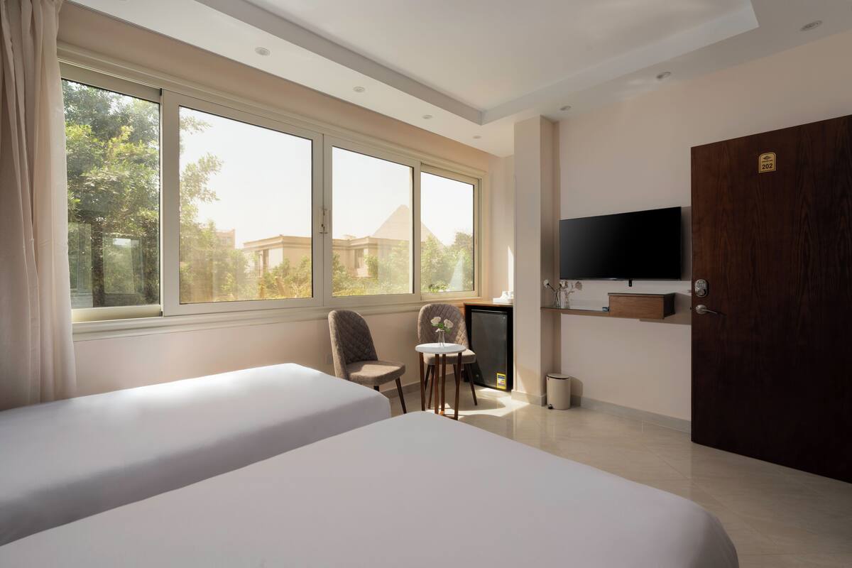 A hotel room with two beds, a desk, a TV, and a large window offering a view of trees and buildings.