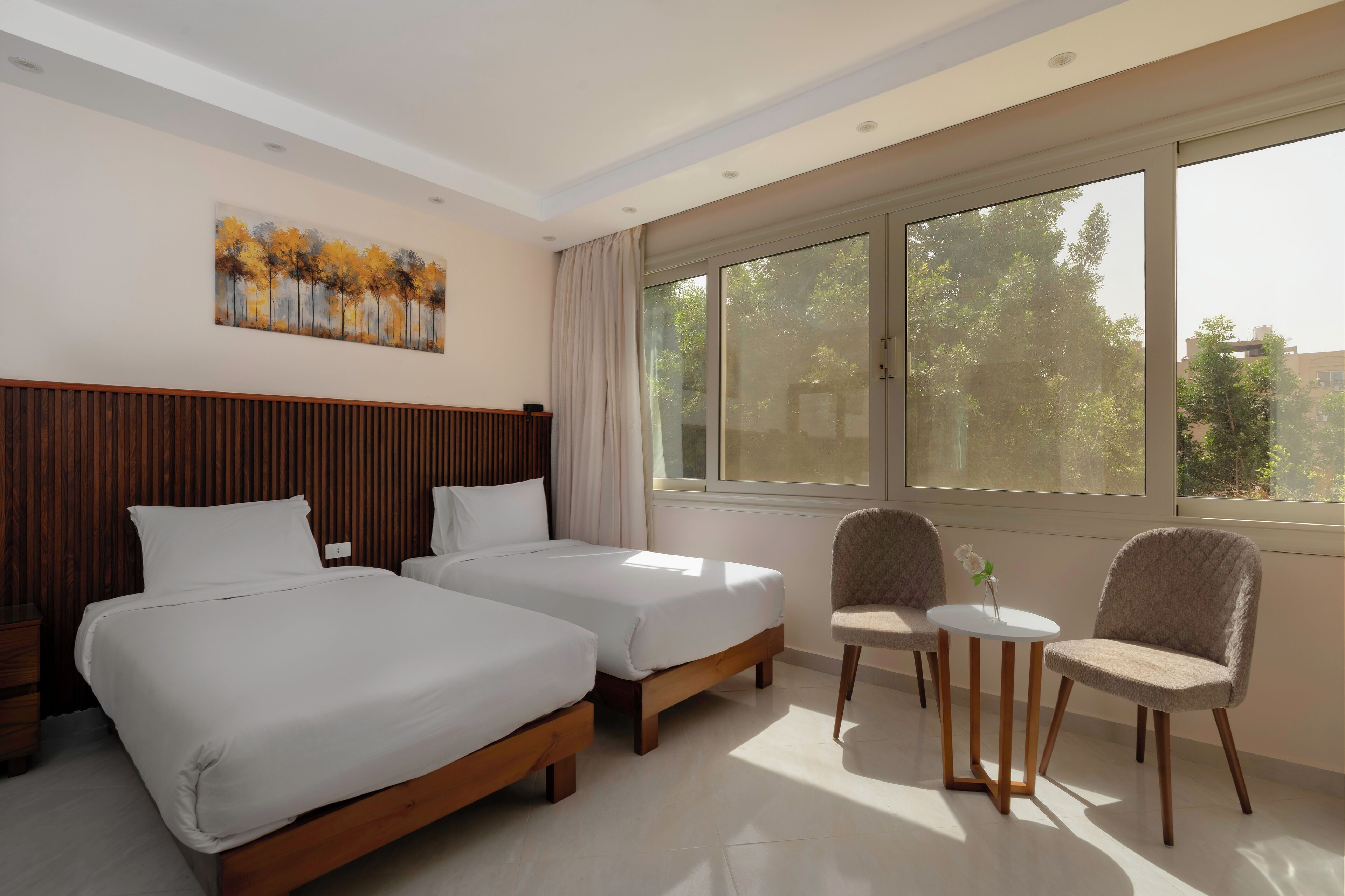 Habitación de hotel con dos camas, una mesita con sillas, un cuadro en la pared y un ventanal con vistas a árboles y edificios.