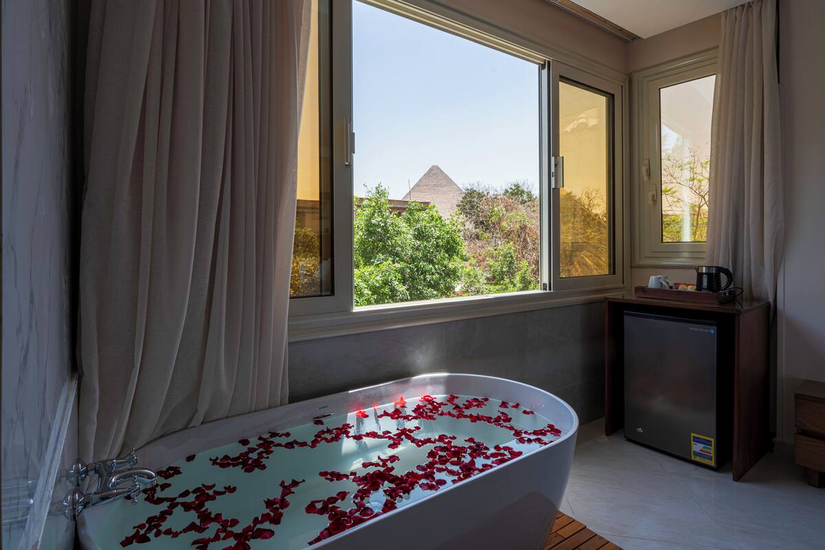 A bathroom with a bathtub filled with rose petals, a coffee machine, and a view of a pyramid and trees outside.