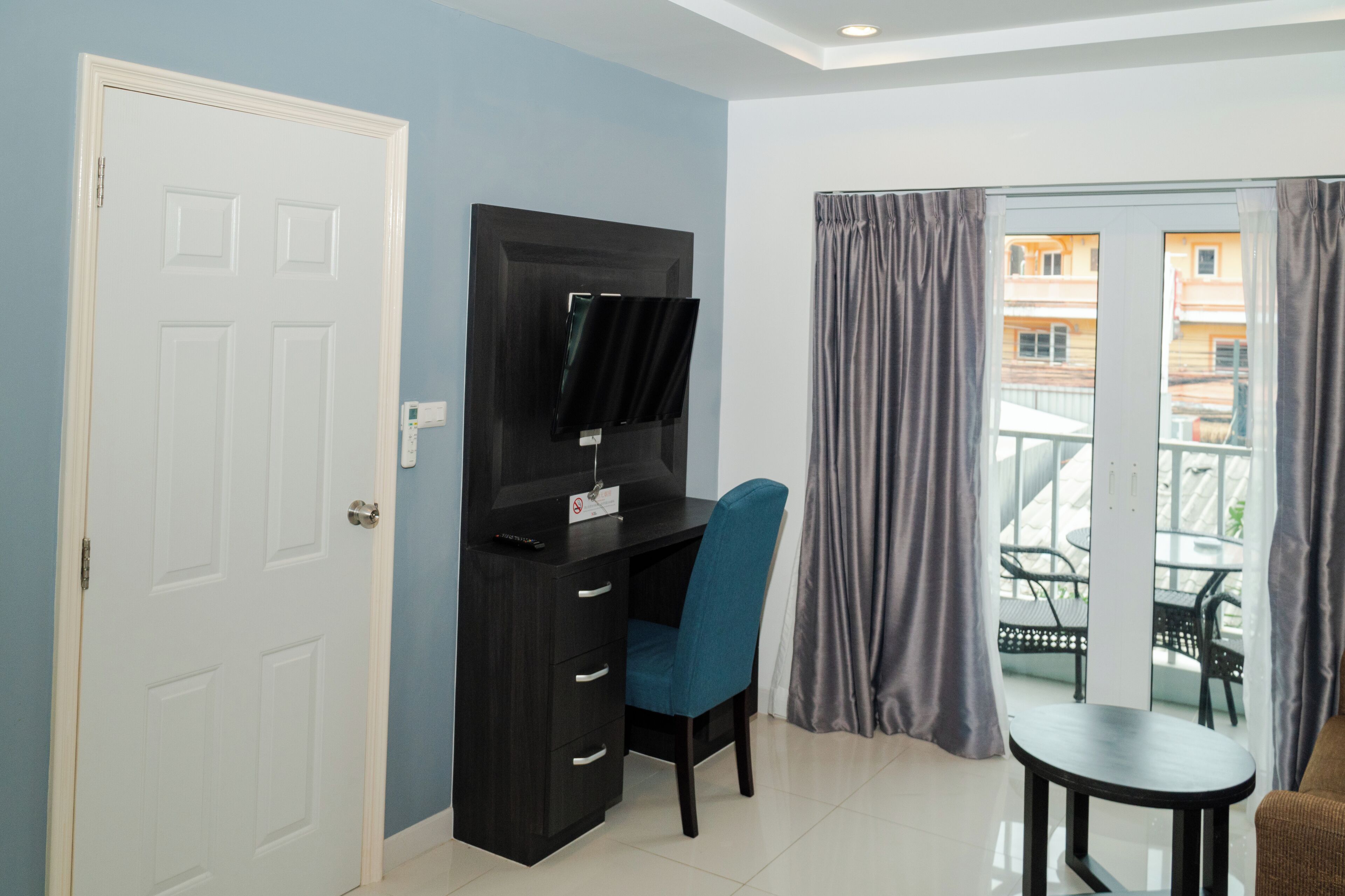 Een kamer met een donker houten bureau, een blauwe stoel, een klein rond tafeltje en een glazen schuifdeur die uitkomt op een balkon met stoelen.