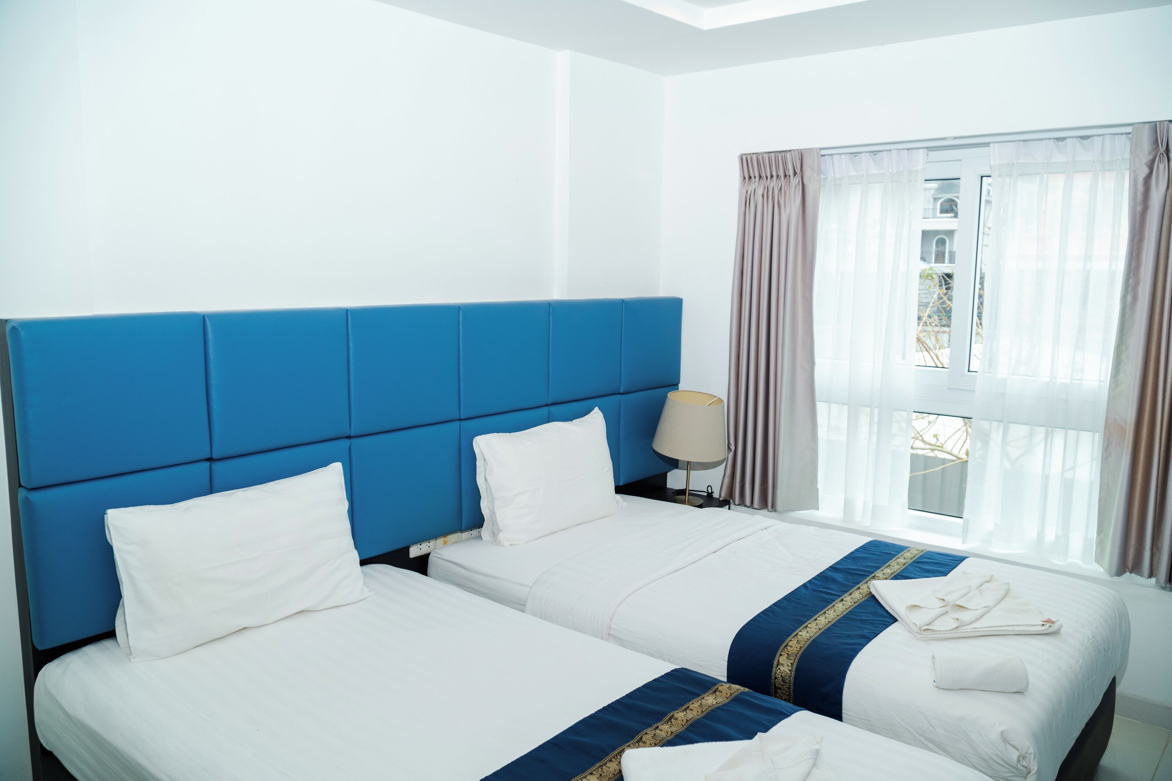 A hotel room with two beds, a blue headboard, white bedding, a window with curtains, and a lamp on the nightstand.