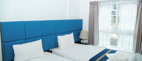 A hotel room with two beds, a blue headboard, white bedding, a window with curtains, and a lamp on the nightstand.