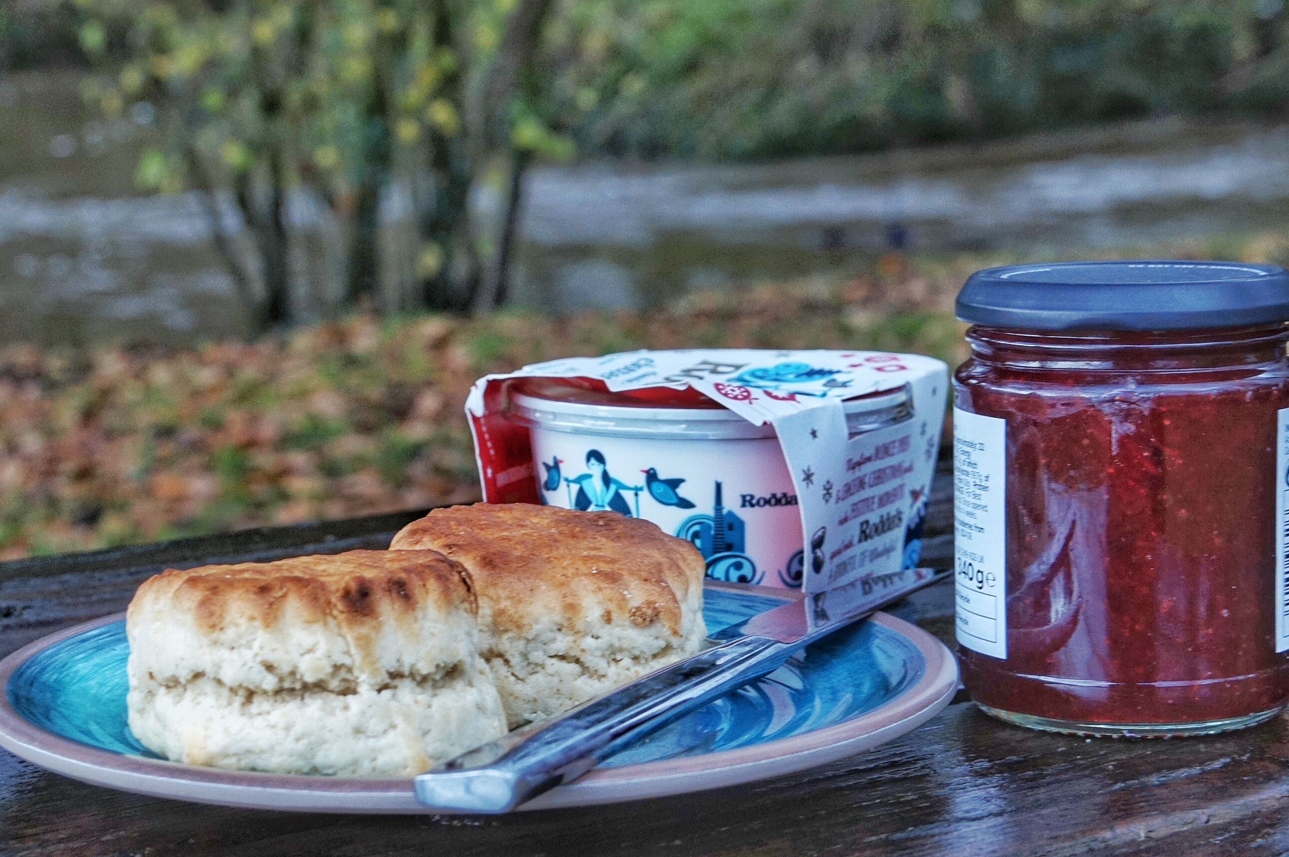 A plate with two biscuits and a jar of jam on a table with a view of trees and a body of water.
