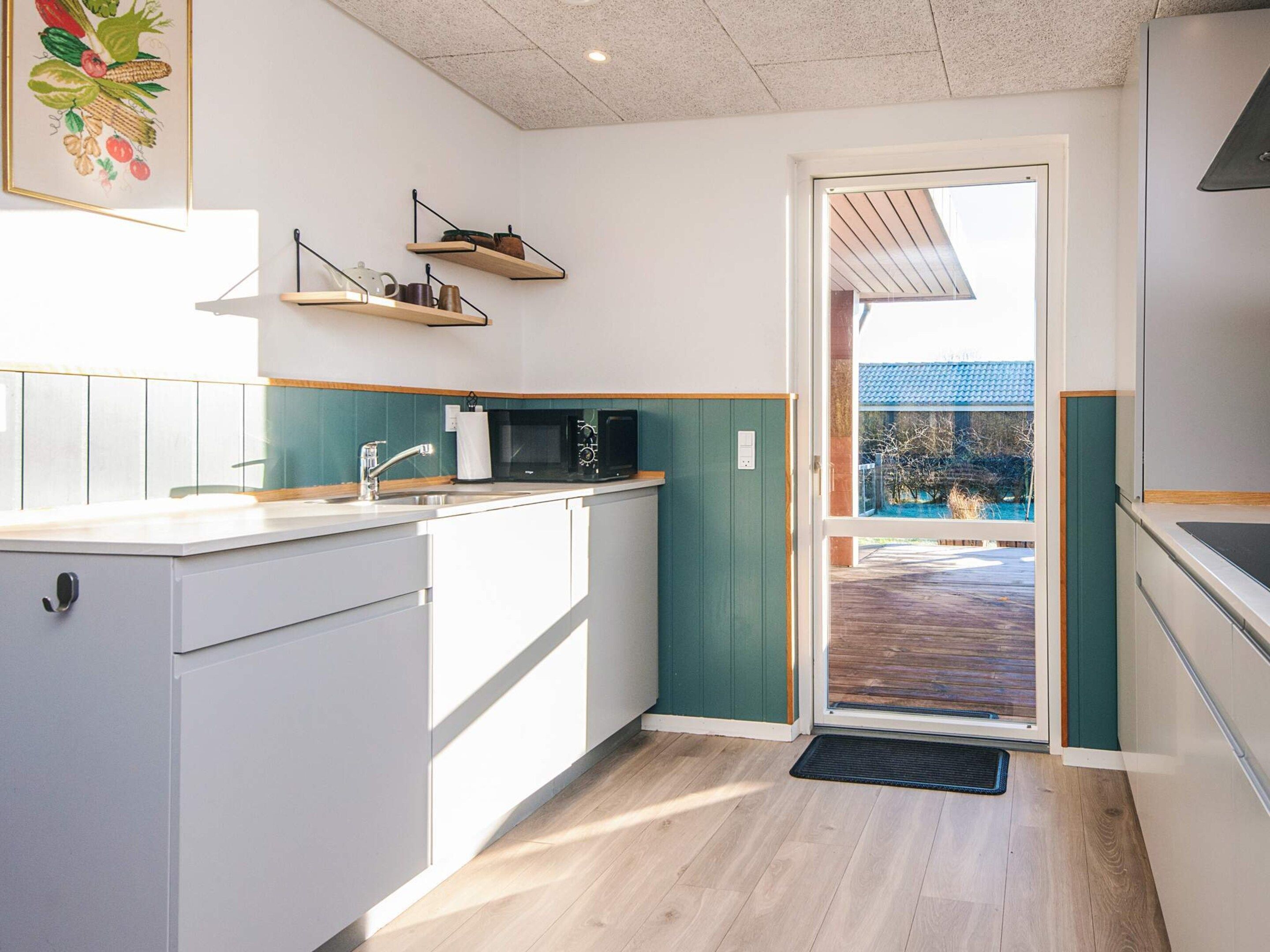 Una cocina moderna con armarios blancos, encimera de madera y una pared verde decorativa. Hay dos estantes colgantes sobre el fregadero y un microondas en la encimera. Una puerta da a un espacio exterior con terraza de madera.