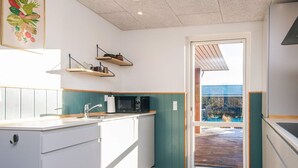 A modern kitchen with white cabinets, a wooden countertop, and a green accent wall. There are two hanging shelves above the sink and a microwave on the counter. A door leads to an outdoor area with a wooden deck. - 5 Star Holiday Home in Tarm (Tarm)