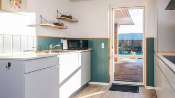 Una cocina moderna con armarios blancos, encimera de madera y una pared verde decorativa. Hay dos estantes colgantes sobre el fregadero y un microondas en la encimera. Una puerta da a un espacio exterior con terraza de madera.