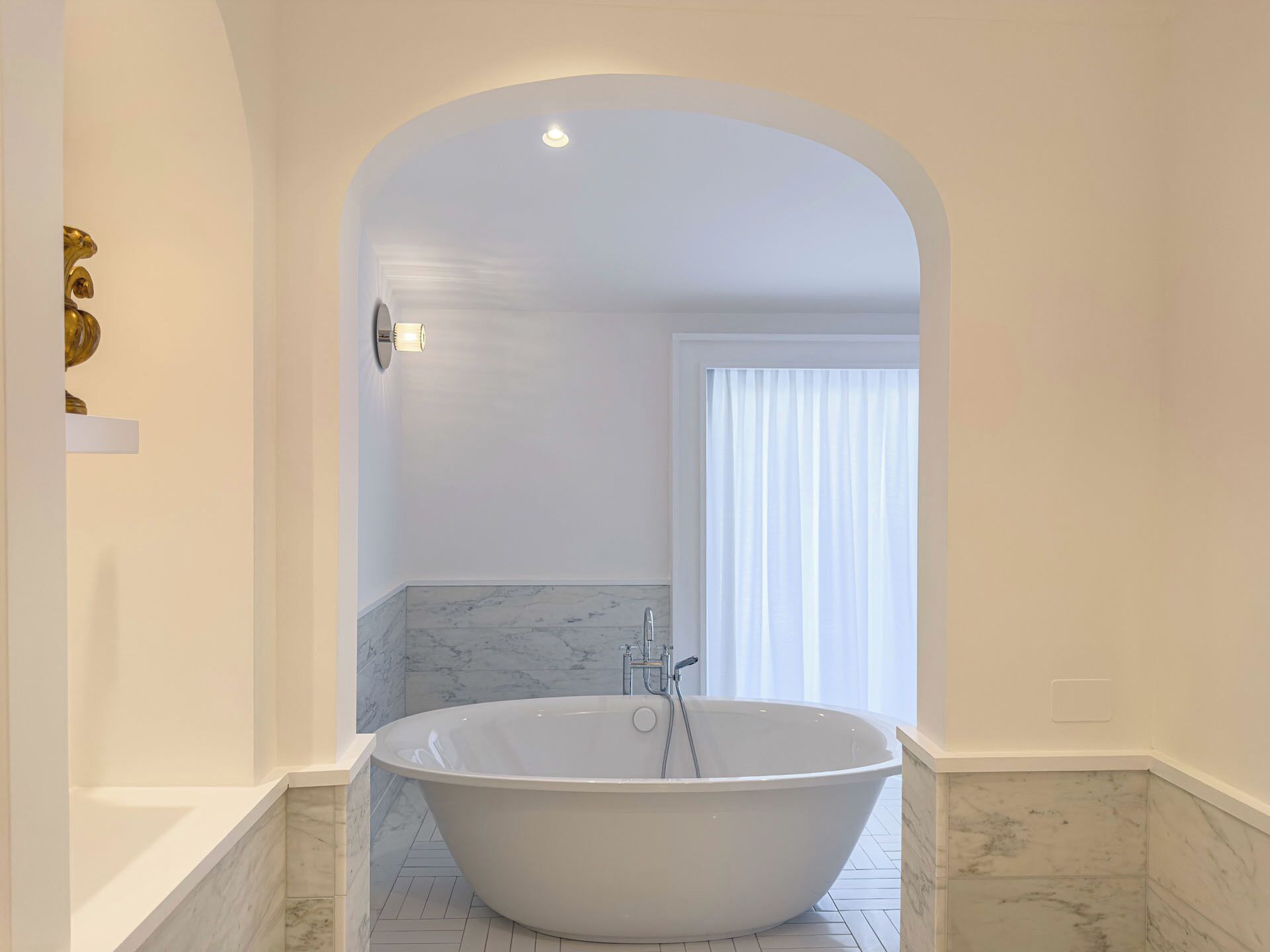 Un baño moderno con una bañera independiente, suelo de mármol y un arco blanco que da a una ventana con cortinas translúcidas.