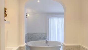 Un baño moderno con una bañera independiente, suelo de mármol y un arco blanco que da a una ventana con cortinas translúcidas.