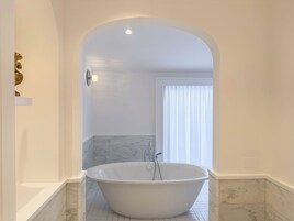 Un baño moderno con una bañera independiente, suelo de mármol y un arco blanco que da a una ventana con cortinas translúcidas.