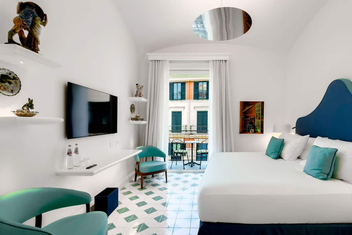 A hotel room with a bed, a TV, a balcony with chairs, and a view of buildings.