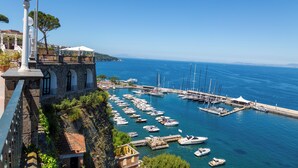A coastal marina with boats docked, a stone building with arched windows, and a clear blue sky.