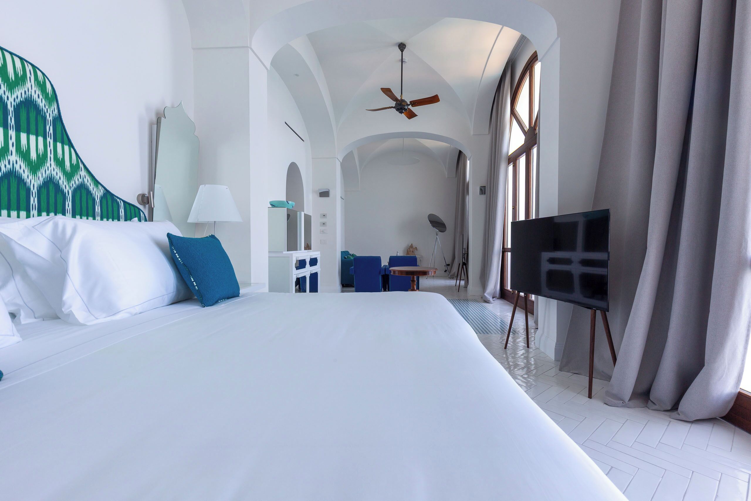 Habitación de hotel con una cama grande, ventilador de techo, televisor sobre un mueble y vista al pasillo.