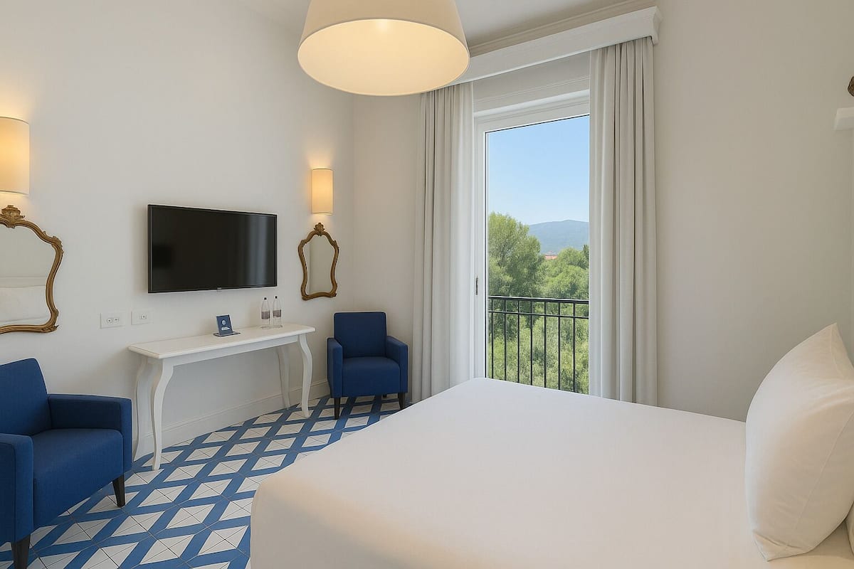 A hotel room with a bed, two blue armchairs, a desk with a flat-screen TV, and a large window with a view of trees and mountains.