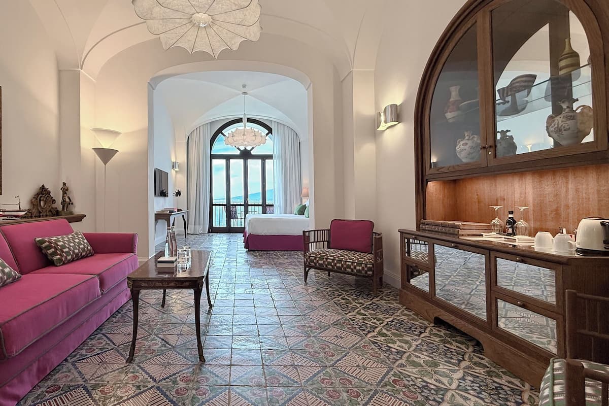 A hotel room with a pink sofa, a wooden side table, a patterned floor, a bed with a view, and a large mirror.