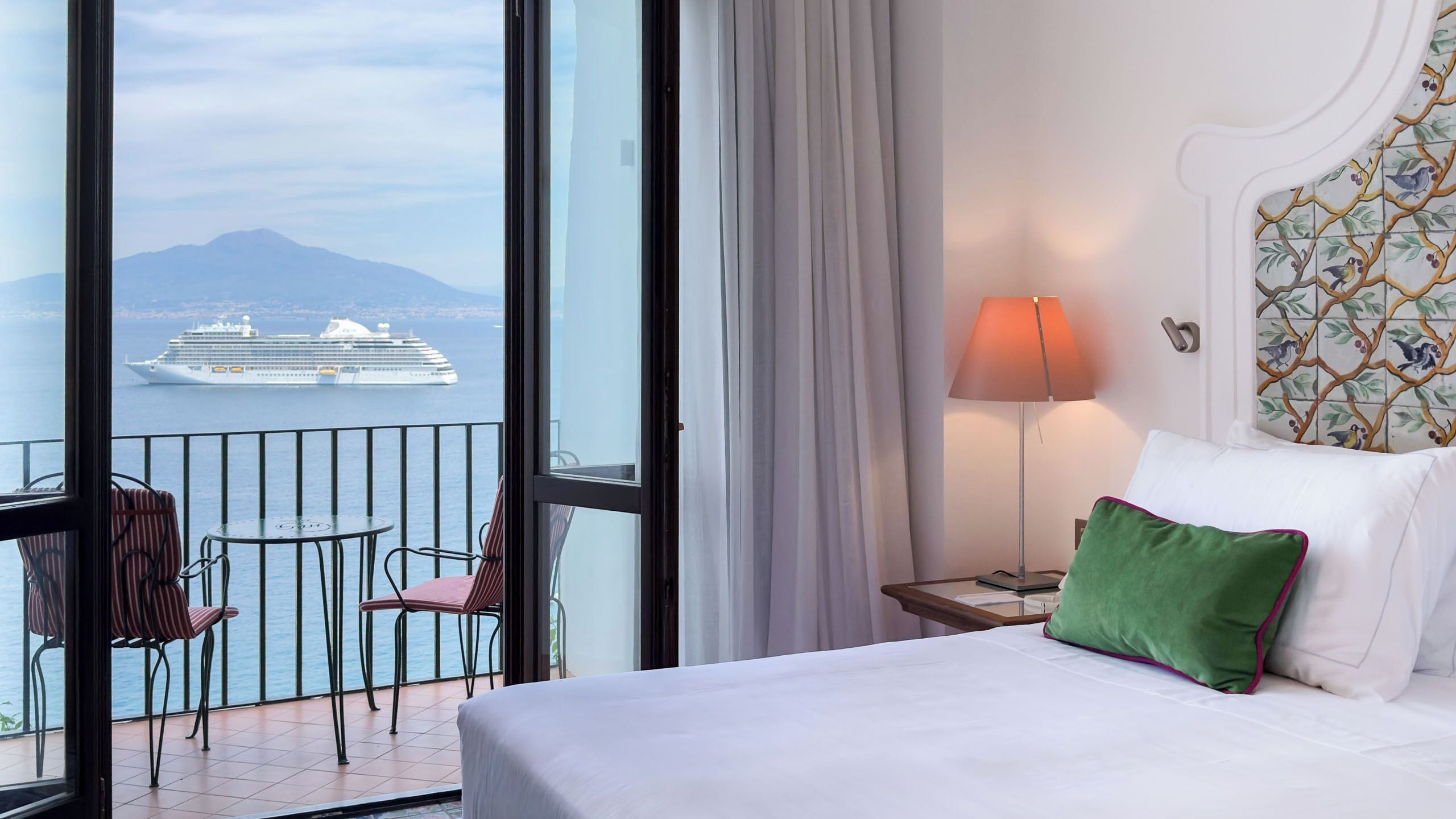 A hotel room with a balcony, a bed with white bedding, a green pillow, a lamp, and a view of a cruise ship.