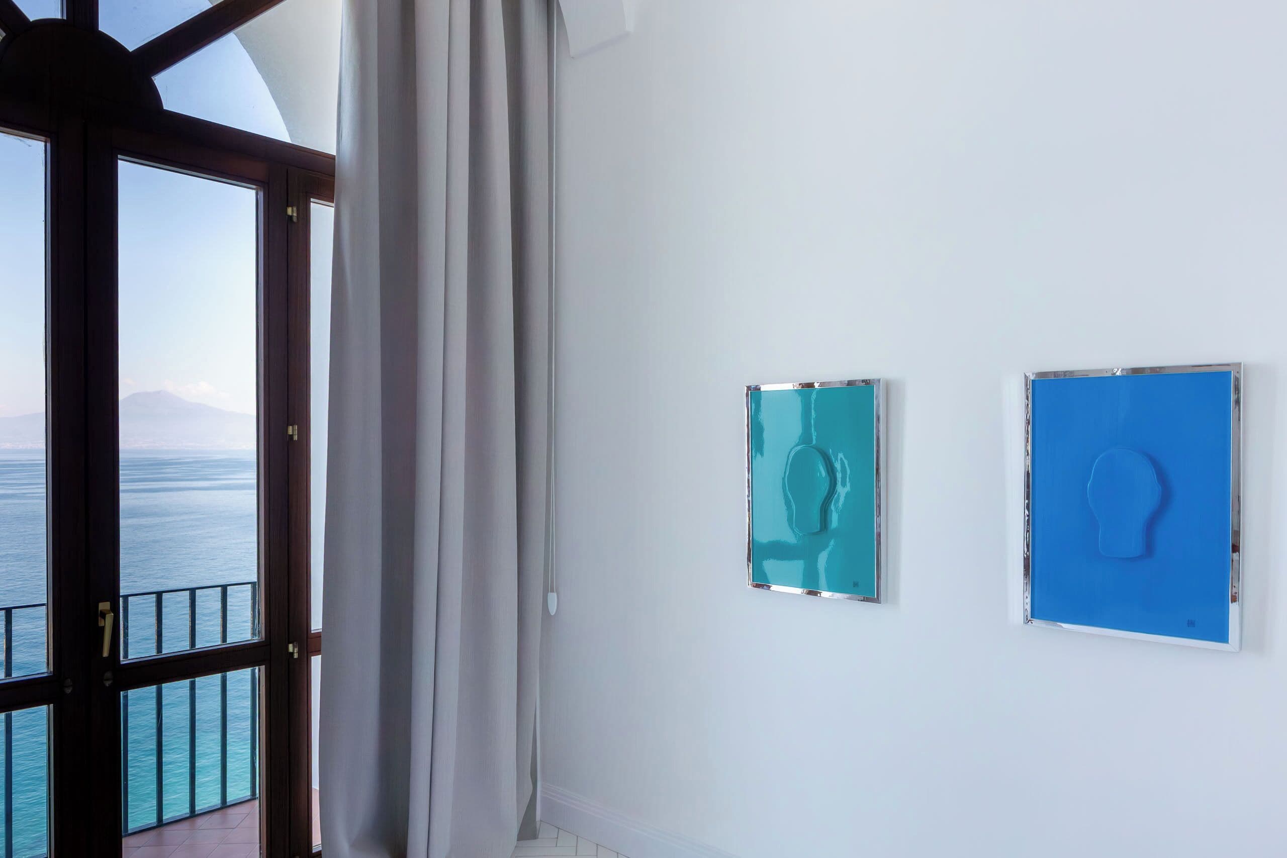 Una habitación con un ventanal grande con vista al mar, una cortina y dos cuadros en la pared.