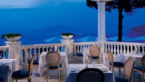 A restaurant with tables set for dining, overlooking a scenic view of the sea and mountains.