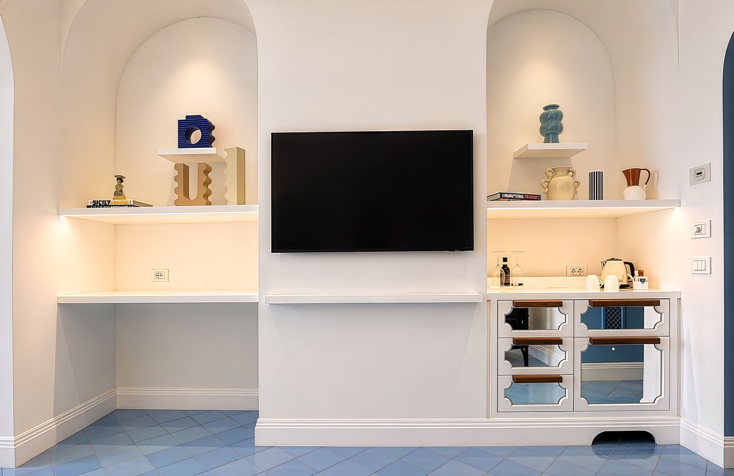 Ein Zimmer mit Wand-Fernseher, weißer Regalanlage mit Dekoration, weißem Schrank mit Glastüren und blauen Fliesen.