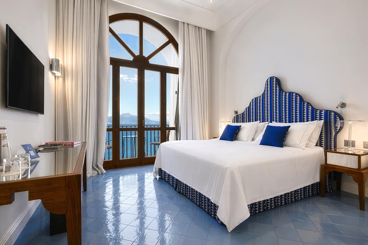 A hotel room with a large bed, a TV, a desk, and a view of the sea.