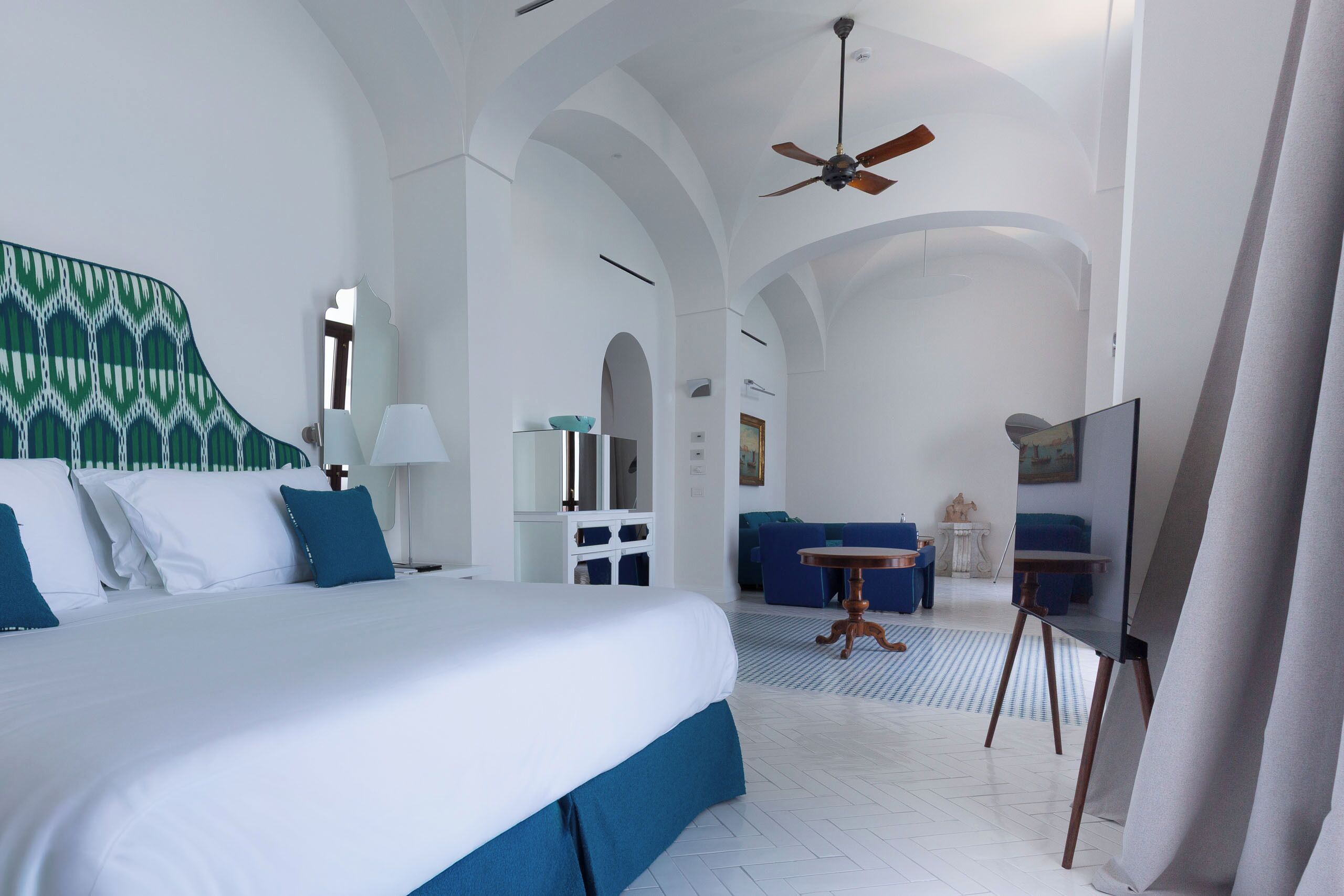 Habitación de hotel con una cama grande, ventilador de techo, escritorio con silla, una mesita y un cuadro en la pared.