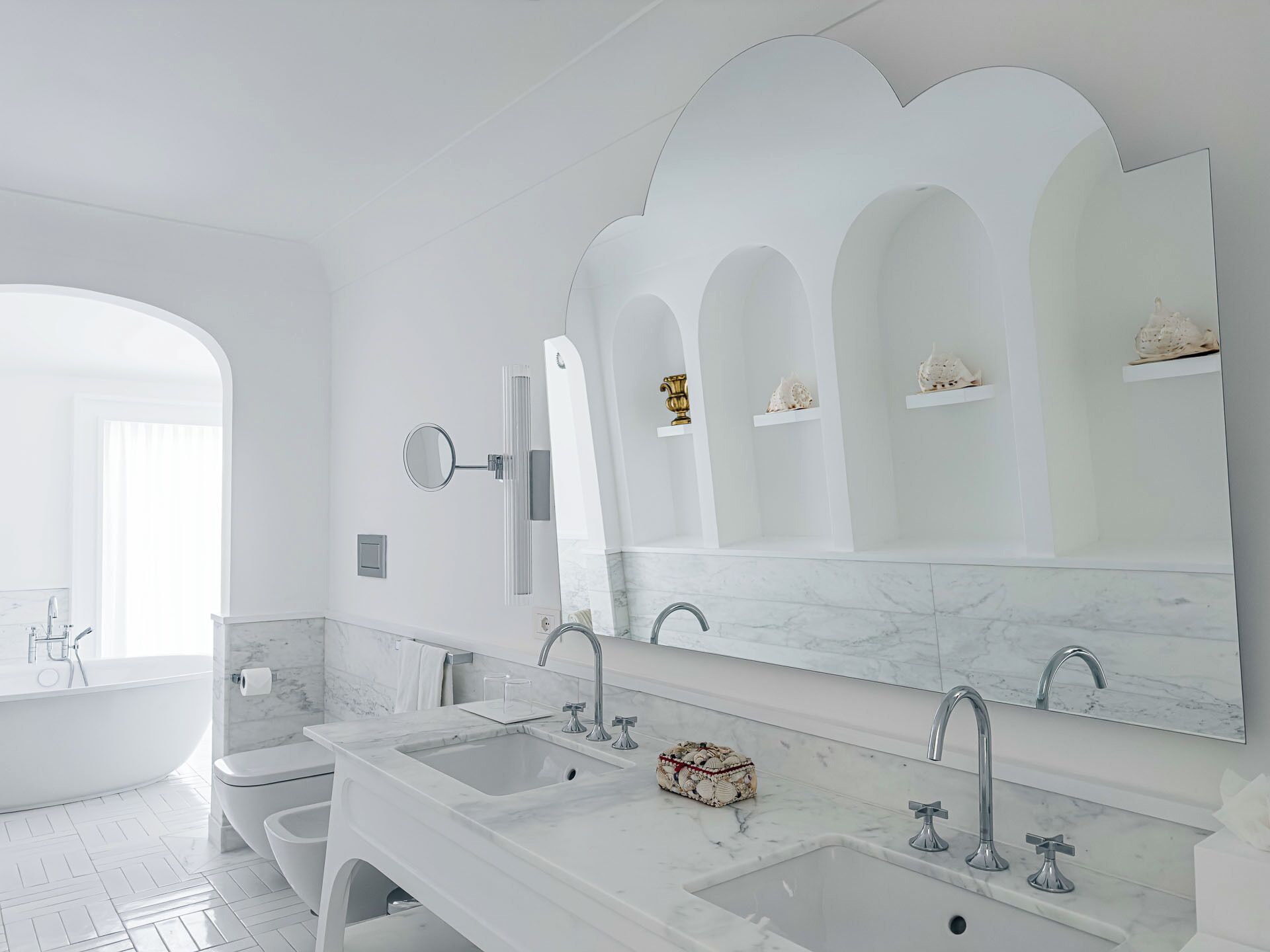 A modern bathroom with a large mirror, two sinks, a bathtub, and decorative items on shelves.