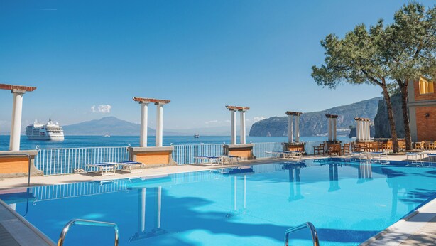 A pool with lounge chairs and umbrellas, a cruise ship in the background, and a mountain view.