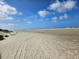 A wide sandy beach with tire tracks, a clear blue sky, and a distant lighthouse.
