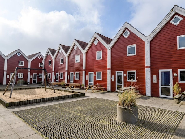 A row of red buildings with white trim, outdoor seating, and a paved area with a central planter. - 4 Person Holiday Home in Fano (Fano)