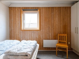 Une chambre avec un mur lambrissé en bois, une fenêtre, un lit recouvert d’une couette blanche, une chaise en bois et un radiateur.
