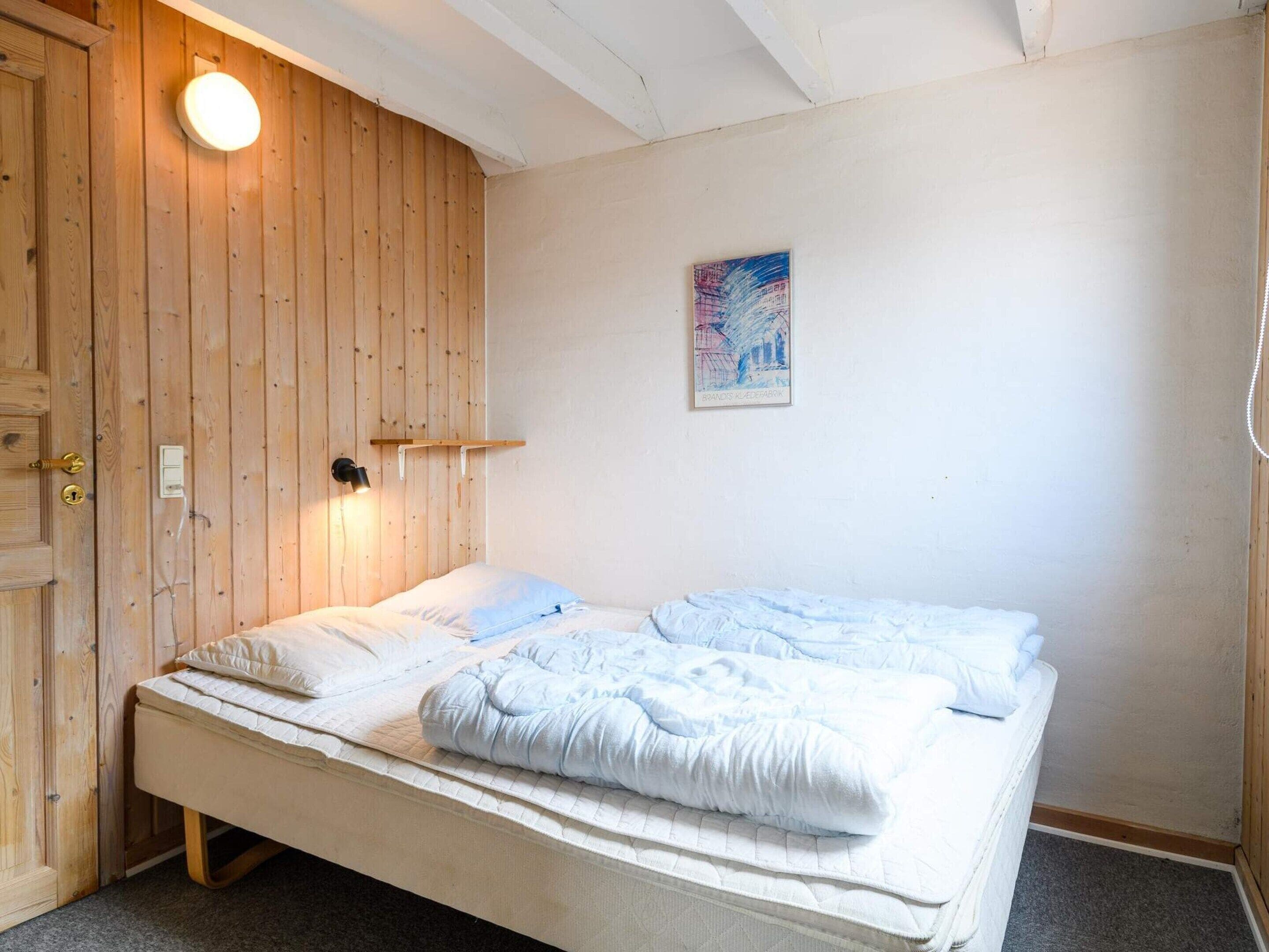 Un lit simple avec un matelas blanc et une literie bleue, une porte en bois, une lampe fixée au mur et un tableau encadré.