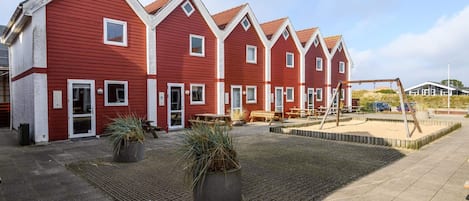 A row of red buildings with white trim, outdoor seating, and a sandy area with a swing set.