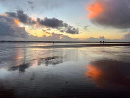 Une plage au sable humide qui reflète le ciel, quelques personnes au loin et une mer calme.