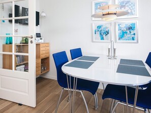 Een kamer met een witte tafel, blauwe stoelen, een boekenkast en kunstwerken in lijsten aan de muur.