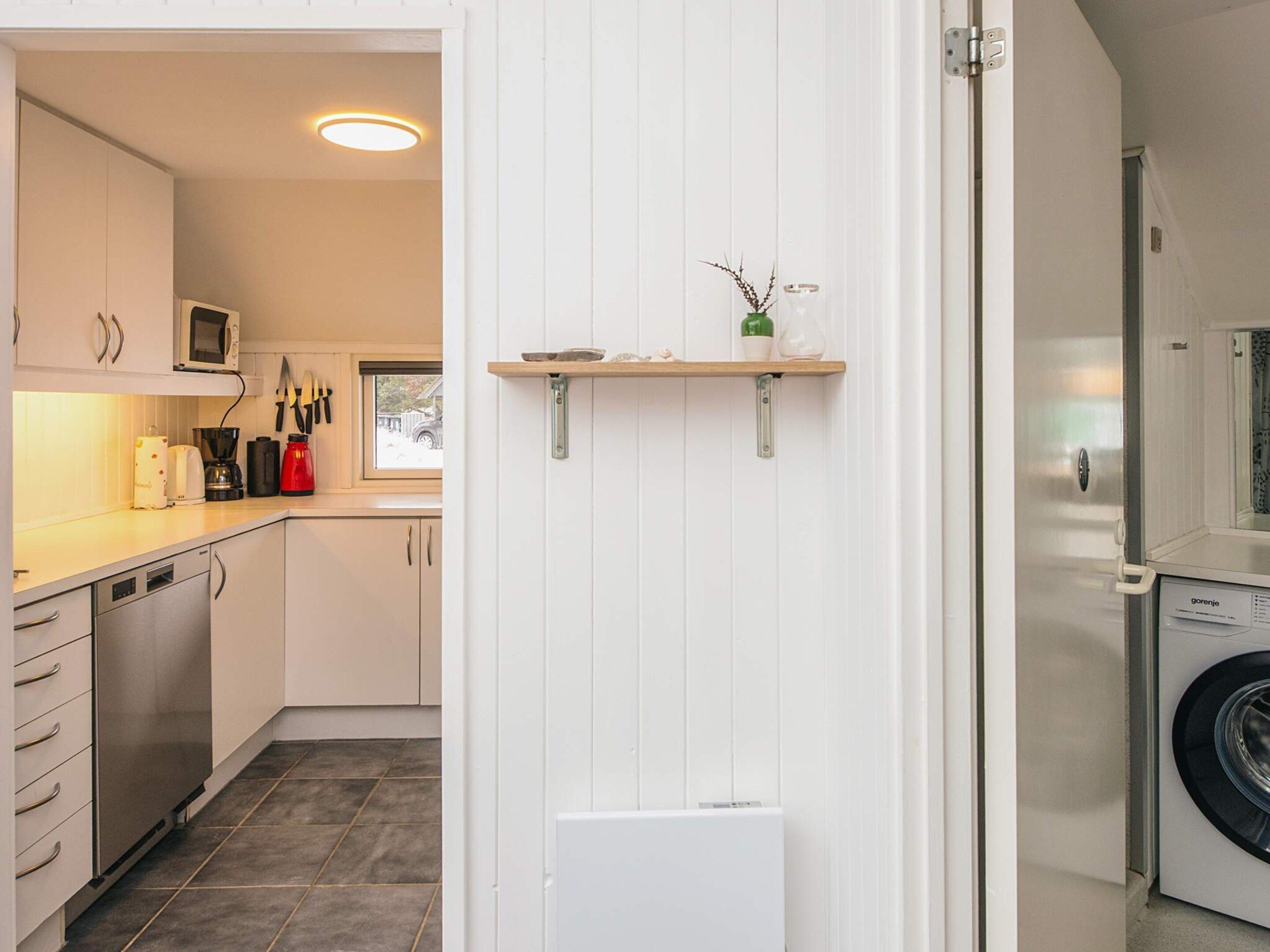 Cocina compacta con armarios blancos, microondas y cafetera. Se aprecia una pequeña estantería con objetos decorativos y una ventana con vistas al exterior.