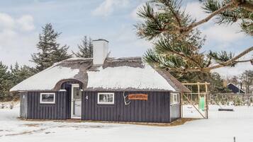 A small, dark wooden cabin with a snow-covered roof and a chimney, surrounded by trees.
