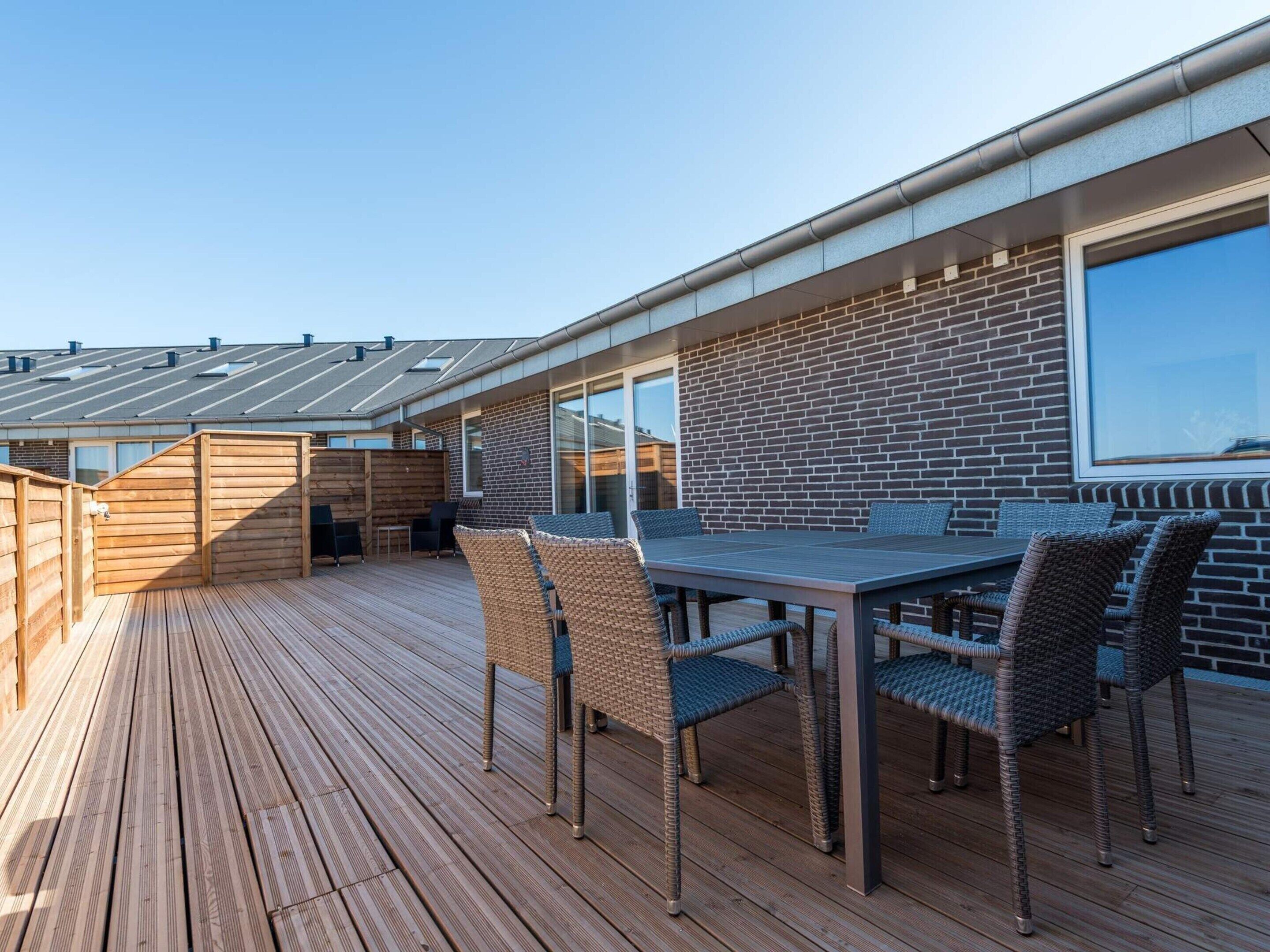 Une terrasse en bois avec une table et des chaises, un mur en briques et une grande fenêtre.