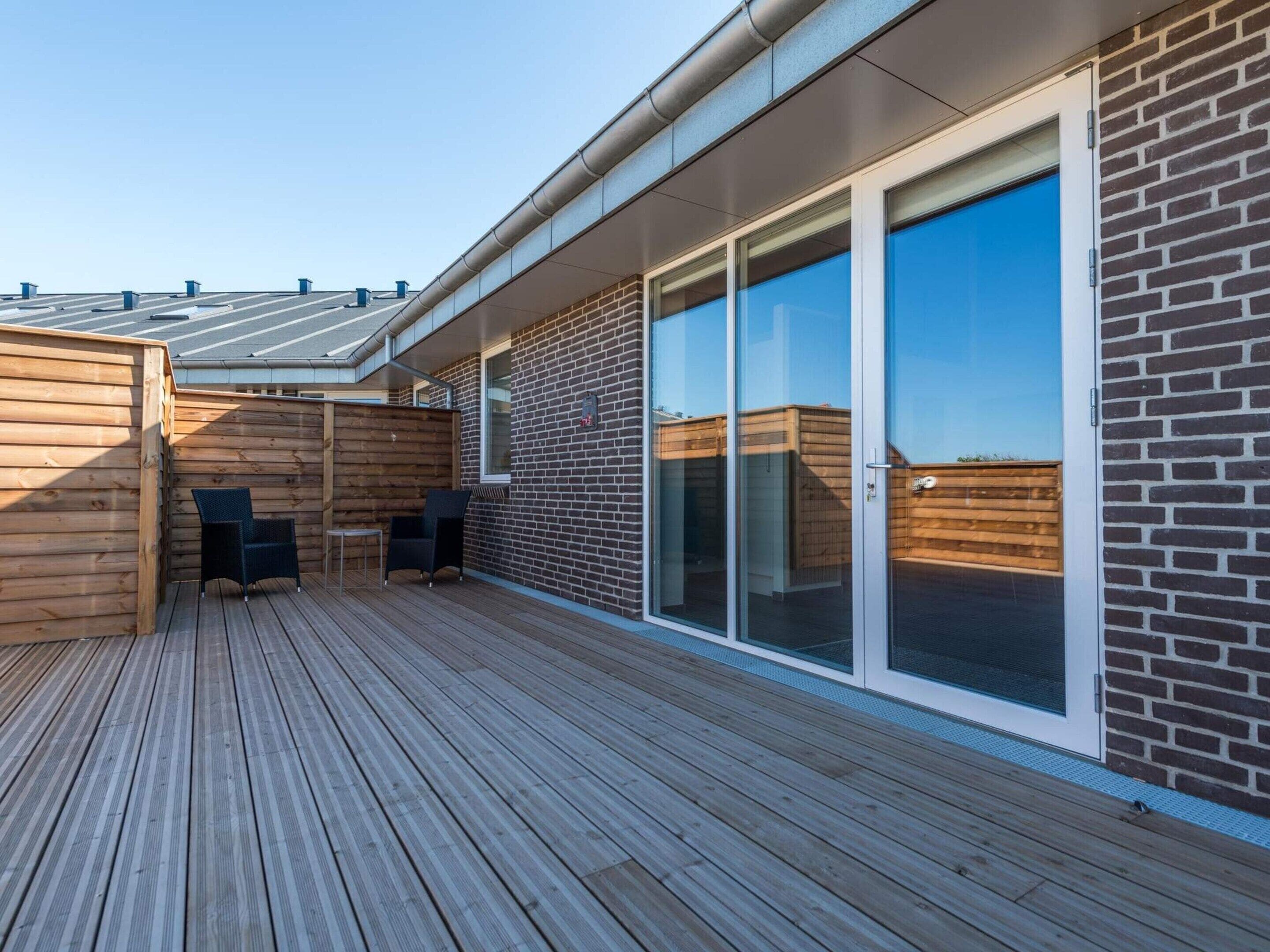 A modern outdoor deck with a wooden floor, two black chairs, and a glass door leading to a covered area.