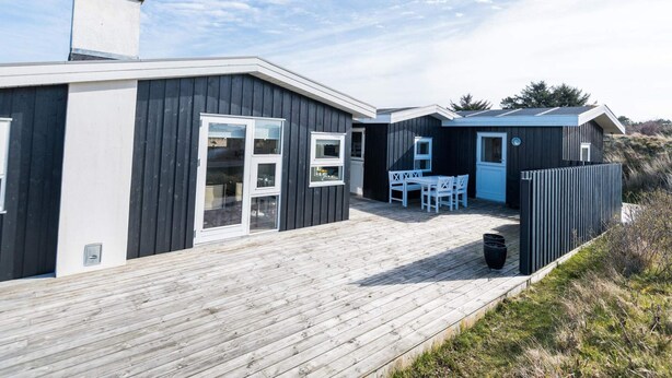 Un bâtiment moderne situé en bord de mer, doté d’une terrasse en bois, de grandes fenêtres et d’un petit espace extérieur aménagé pour s’asseoir.