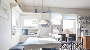 Un espace repas avec une table blanche, des bancs et des chaises. Il y a des fenêtres donnant sur l’extérieur.