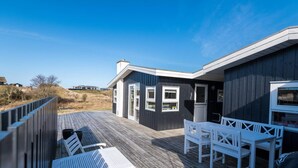 A wooden deck with white furniture, a building with large windows, and a clear sky. - 4 Person Holiday Home in Fano (Fano)