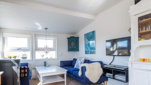 Un salon moderne avec un canapé bleu, une table basse blanche, un téléviseur à écran plat et un coin cuisine doté d’un meuble blanc.