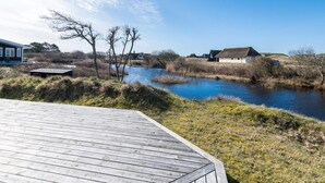 Une propriété située au bord de la rivière, dotée d’une terrasse en bois, d’une maison à toit de chaume et d’un bâtiment moderne en arrière-plan.