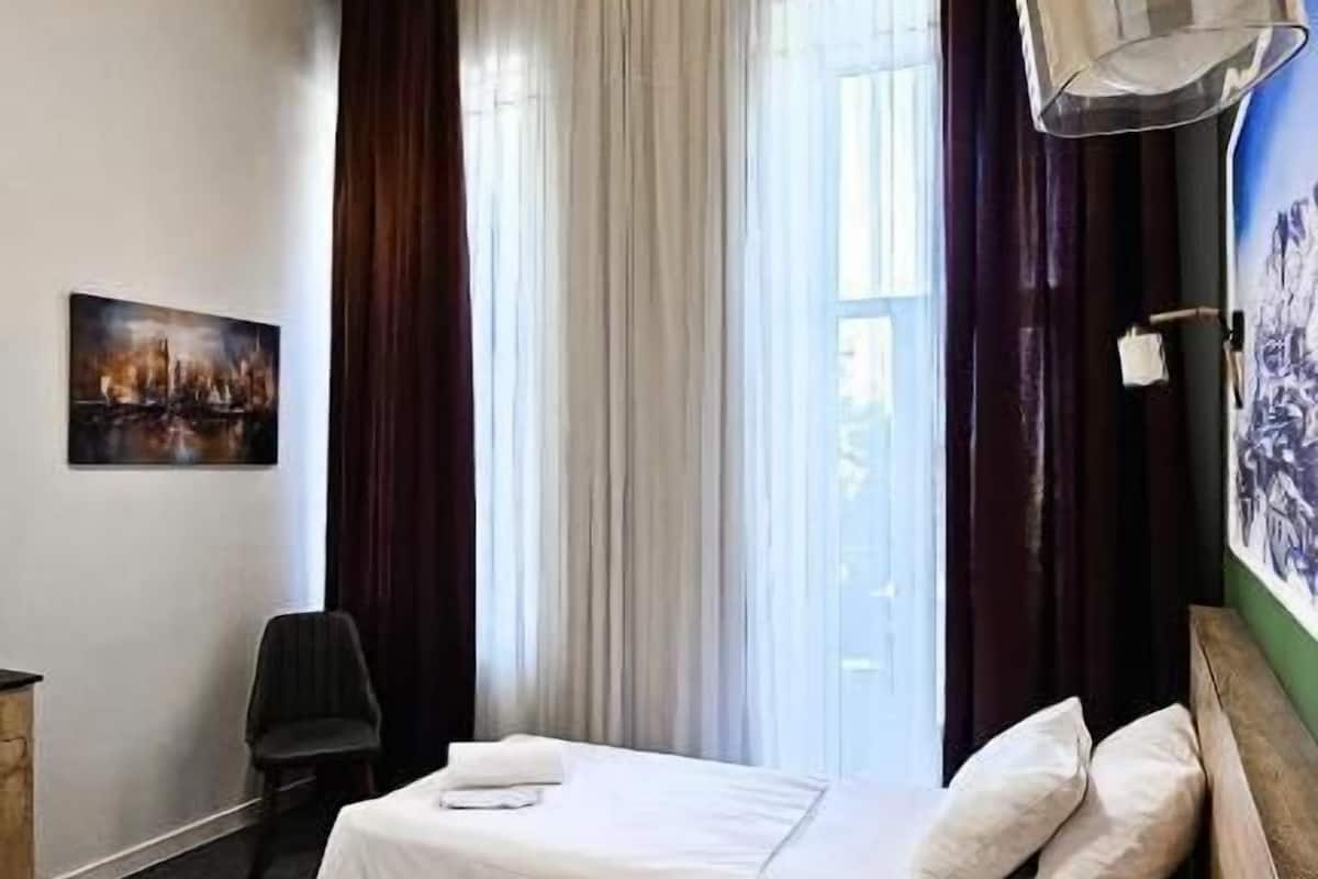 A hotel room with two beds, a large window with curtains, a painting on the wall, and a ceiling light fixture.